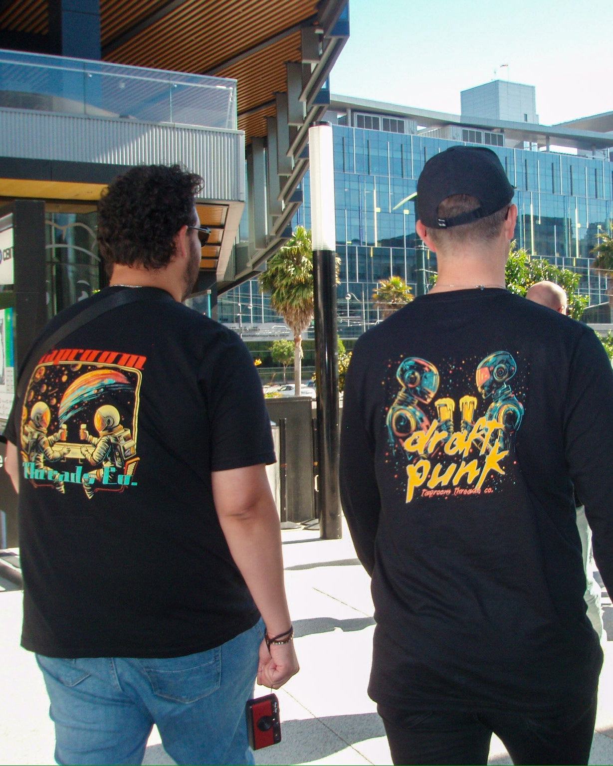 Two people wearing black t-shirts with colorful graphics and text, standing outdoors in an urban setting.
