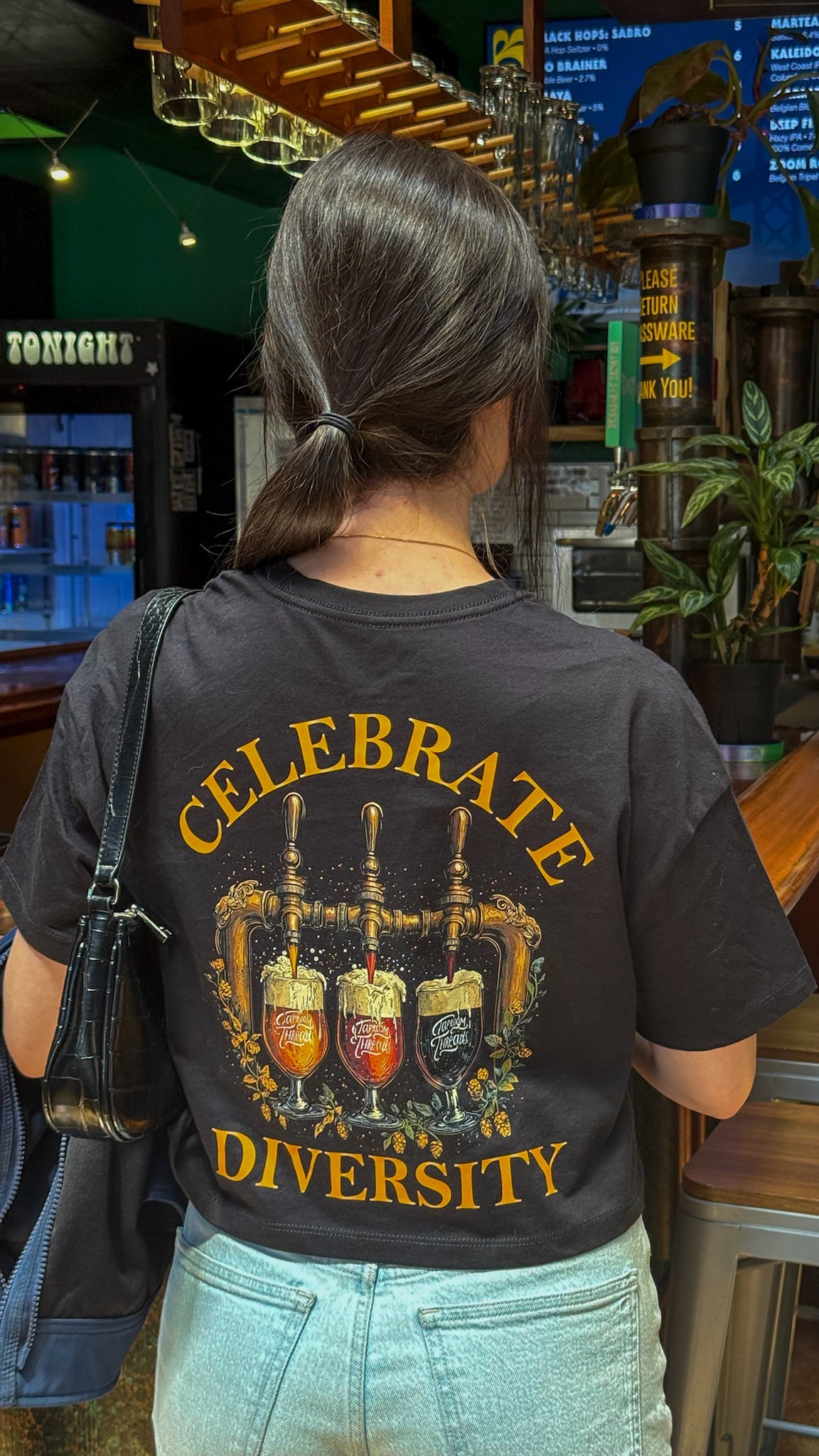 Person wearing a black t-shirt with 'Celebrate Diversity' design in a bar setting